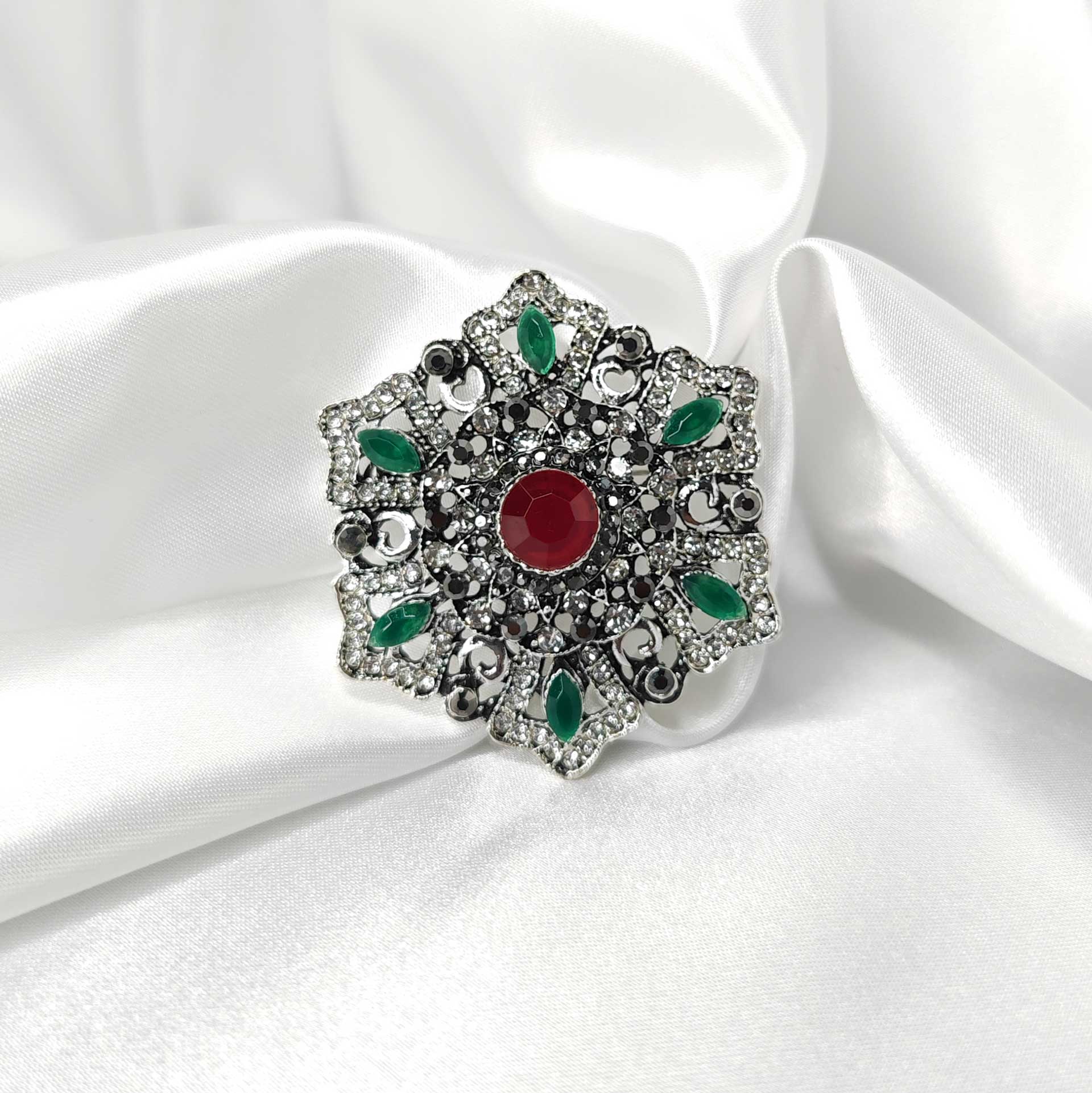 Photo principale de la broche style baroque a la forme étoilée florale ornée d'un strass rouge centrale, 6 de couleurs vertes a l'extrémité des branches et plusieurs de couleurs cristal.