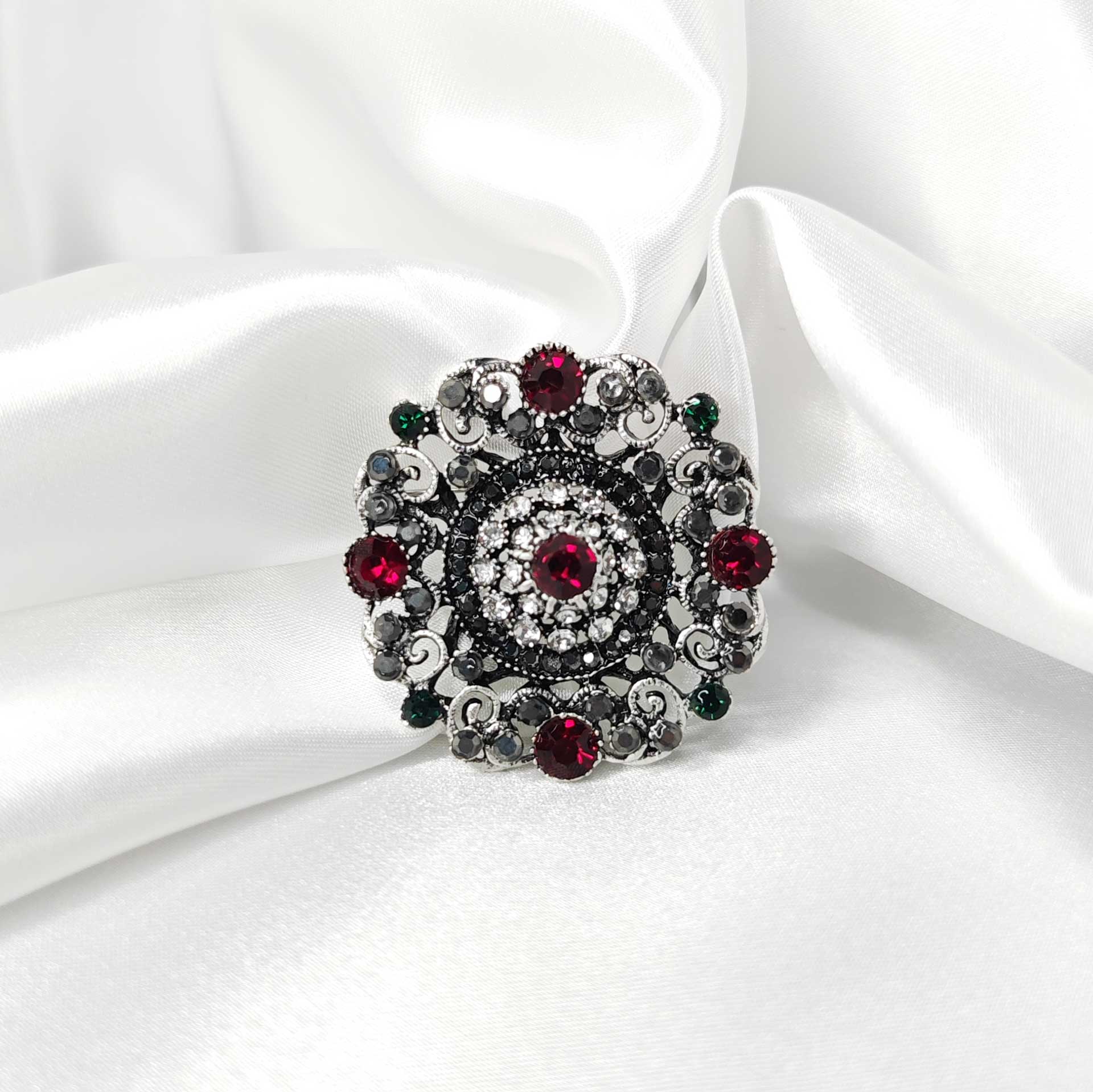 Photo principale de la broche style baroque en forme de rosace mandala ornée de 5 strass rouges cardinaux et des éclats de strass verts, noir et blanc.