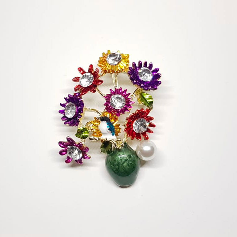 Broche dorée en forme de pot de fleur vert et marguerites multicolores ainsi qu'un oiseau sur des marguerites.