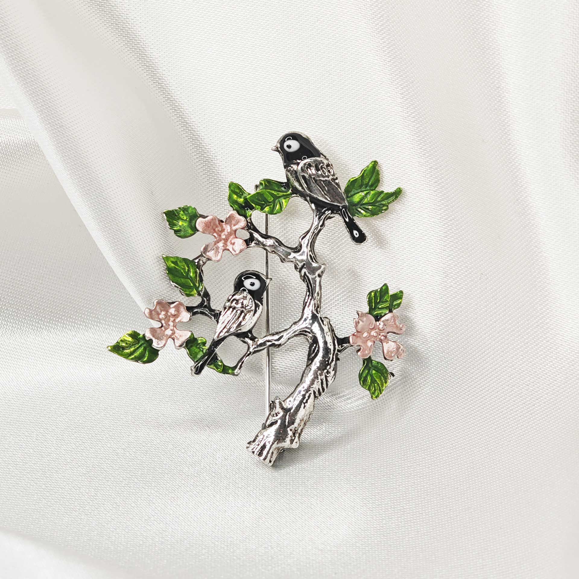 Photo de face de la broche argentée représentant 2 oiseaux posé sur un arbre au feuillage vert et fleurs roses