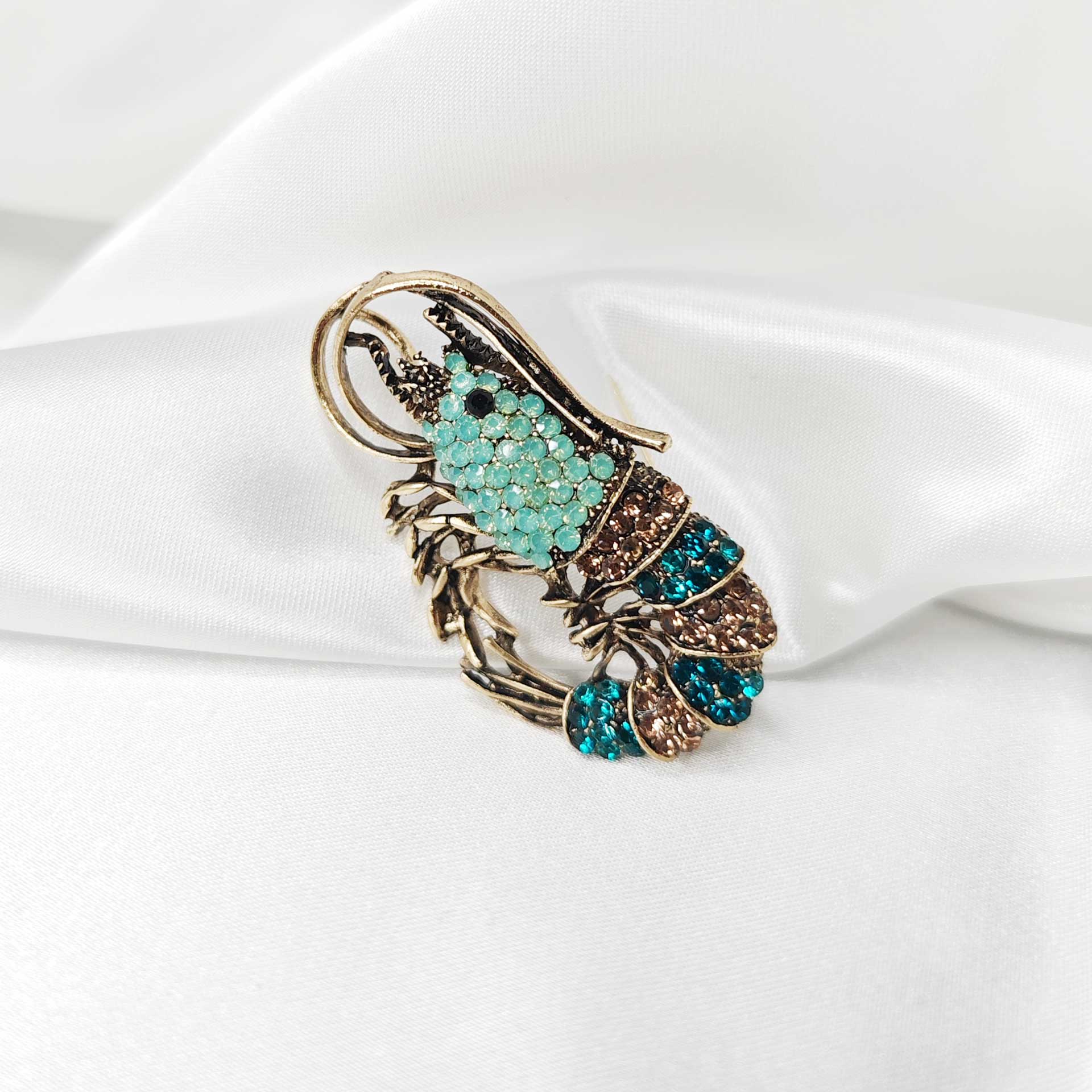 Photo principale de la Broche fantaisie en forme de crevette, ornée de strass bleus et turquoise scintillants, avec détails dorés et antennes fines.