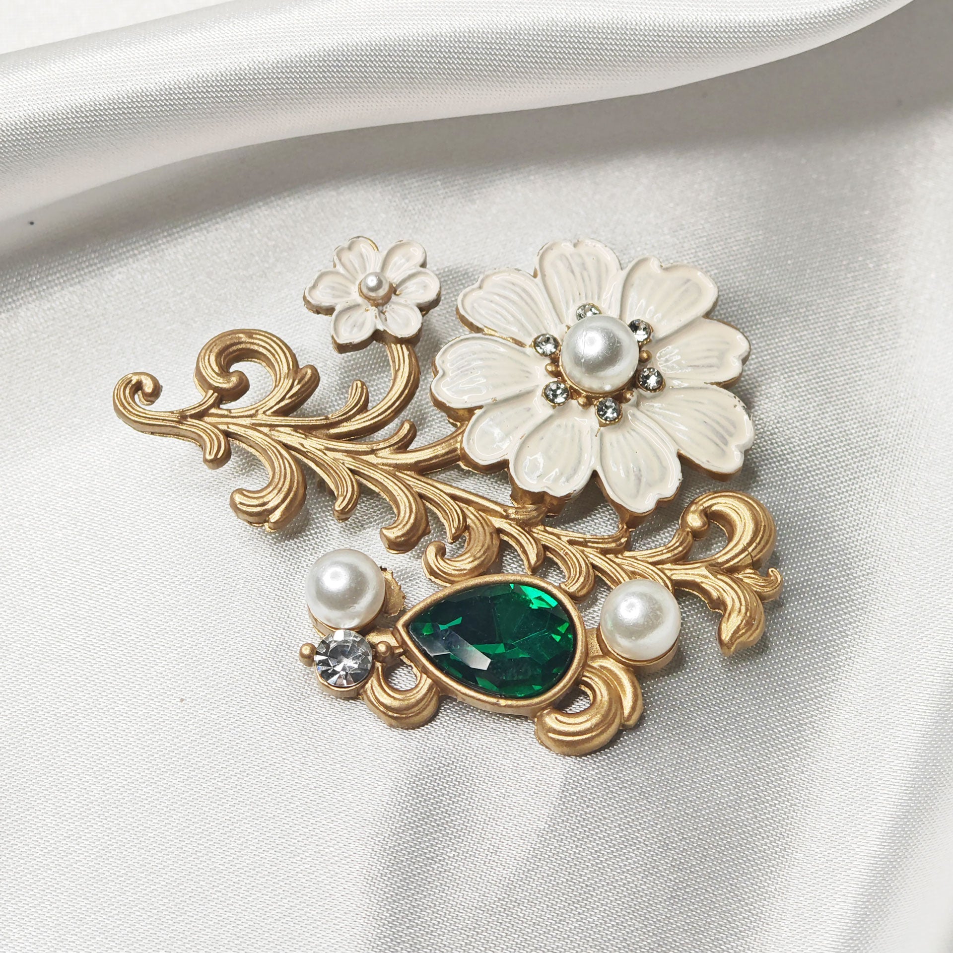 Broche fleur de cerisier baroque dorée avec perle et cristal vert émeraude sur fond satin blanc